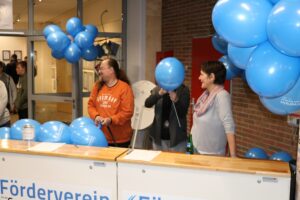 Drei Personen stehen an einem Tisch mit blauen Luftballons und einem Schild des Fördervereins.
