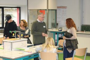 Personen in einem Klassenzimmer interagieren. Tische und Lehrmaterialien sind sichtbar.