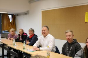Gesprächsrunde mit mehreren Personen an einem Tisch in einem Klassenraum.