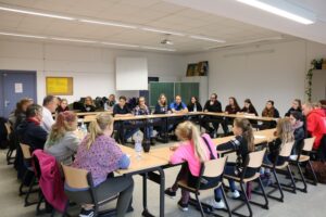 Gruppengespräch in einem Klassenzimmer mit Schülern und Lehrern an Tischreihe.