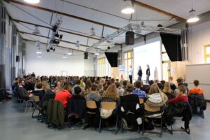 Große Versammlung in einem Schulgebäude mit Schülern und Vortragenden auf der Bühne.