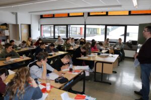 Schüler sitzen in einem Klassenraum, einige schreiben, andere hören zu.