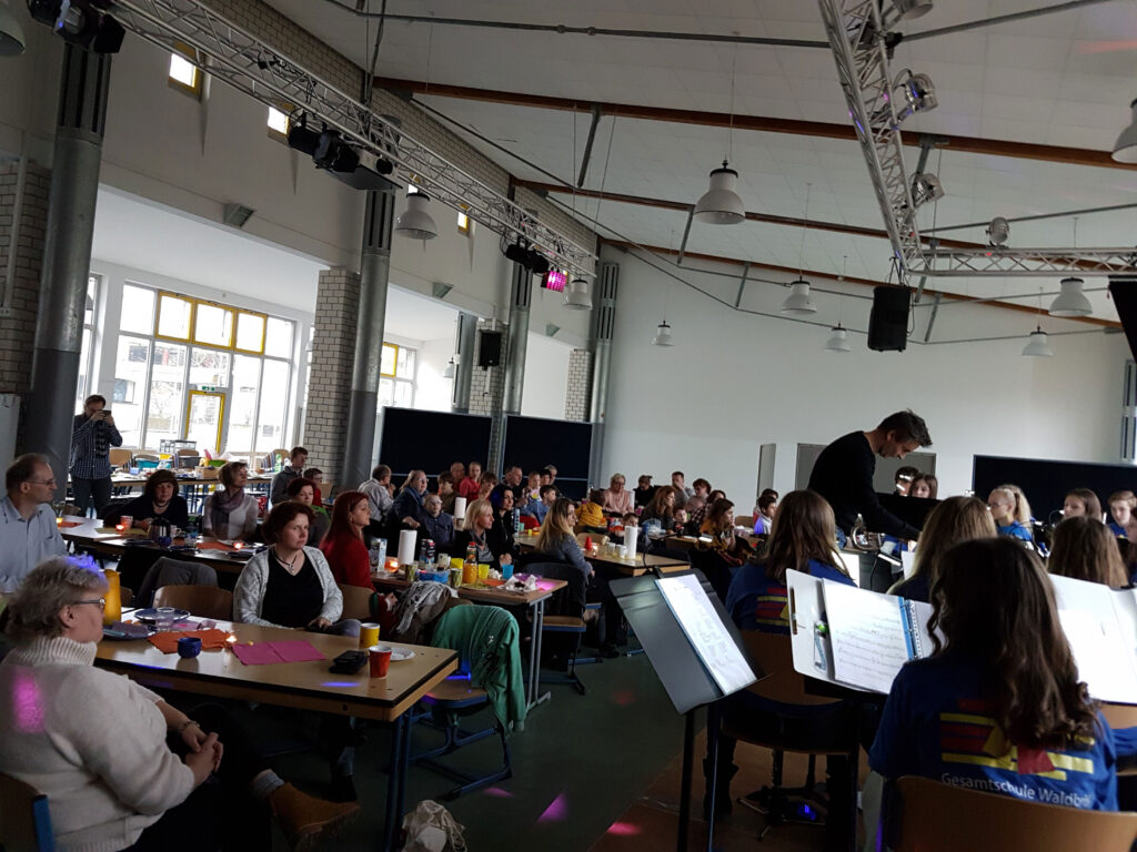 Menschen sitzen an Tischen in einem großen Raum, eine Band spielt vorne.