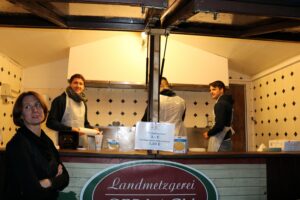 Frau steht vor einem Verkaufsstand mit Mitarbeitern in Schürzen.