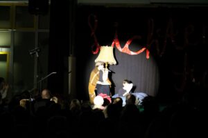 Theateraufführung mit Darstellern auf der Bühne und Publikum im Hintergrund.