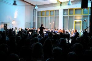 Konzertaufführung mit vielen Zuschauern im Hintergrund und einem Orchester auf der Bühne.