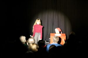 Mädchen auf der Bühne mit Mikrofon, im Hintergrund ein Puppenspieler.