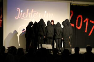 Gruppe von Personen in dunkler Kleidung mit Kapuzen auf einer Bühne.