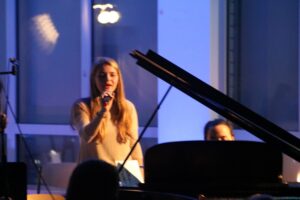 Eine Sängerin tritt vor einem Klavier auf, im Hintergrund ein Musiker.