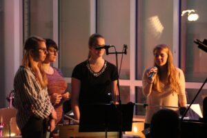 Vier Sängerinnen stehen am Mikrofon und singen in einem beleuchteten Raum.