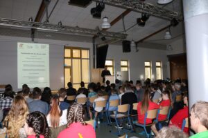 Vortrag in einem großen Saal mit Schülern, die aufmerksam zuhören.