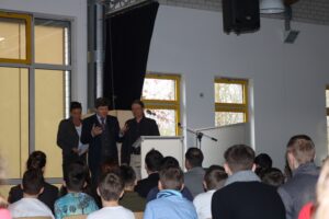 Zuhörer in Aula während einer Ansprache von drei Rednern.