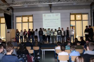 Schüler stehen auf einer Bühne, Publikum sitzt im Vordergrund.