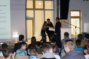 Preisverleihung auf der Bühne mit Schülern und Lehrern. Zuschauer schauen gespannt zu.