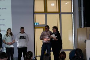 Gruppenvorstellung auf einer Schulveranstaltung mit mehreren Personen auf der Bühne.