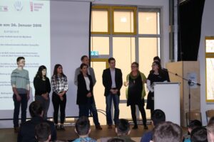 Gruppenvorstellung von Personen auf einer Bühne. Im Hintergrund eine Präsentation.
