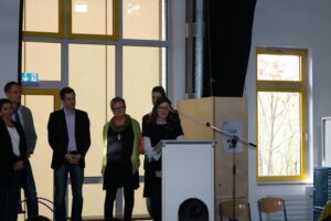 Gruppenansprache vor Publikum in einem hellen Raum mit großen Fenstern.