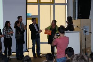 Preisverleihung auf einer Bühne, umgeben von applaudierenden Personen.