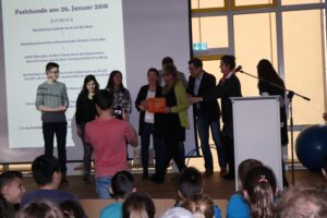 Verleihung von Urkunden an Schüler auf einer Bühne, Publikum im Hintergrund.
