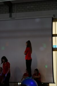 Ein Mädchen steht auf einem Gymnastikball, umgeben von anderen Personen in roten T-Shirts.