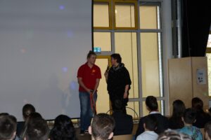 Schüler und Lehrerin auf der Bühne während einer Präsentation. Zuschauer im Vordergrund.
