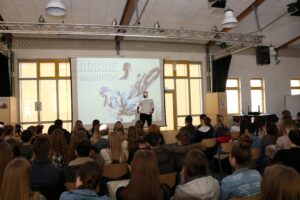 Vortrag in einem Schulraum mit Schülern und Präsentation im Hintergrund.