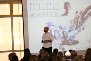 Ein Mann spricht vor einem Publikum, Hintergrund mit Bildern von Robbie Maddison.