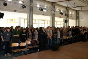Vieler Zuschauer stehen in einem Saal mit Sitzbänken.