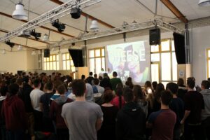 Schüler stehen in einer Aula und hören einer Präsentation zu.