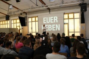 Ein Vortrag in der Aula mit Schülern, der Bildschirm zeigt "EUER LEBEN".
