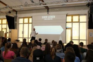 Ein Redner steht vor einer Gruppe von Schülern in einem Vortragsraum.
