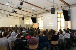 Schüler sitzen in einem großen Raum und hören einem Vortrag zu.