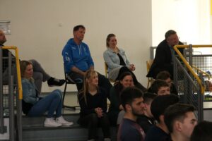 Eine Gruppe von Menschen sitzt auf einer Tribüne in einer Sporthalle.