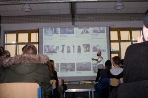 Vortragender steht vor einer Leinwand mit Bildschirminhalt. Zuschauer im Vordergrund.