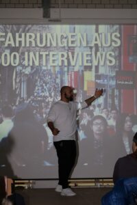 Ein Vortragender steht vor einer Leinwand mit Präsentation über Interviews.