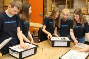 Sechs Schüler stehen um einen Tisch und arbeiten an Projekten mit Materialien in Kisten.
