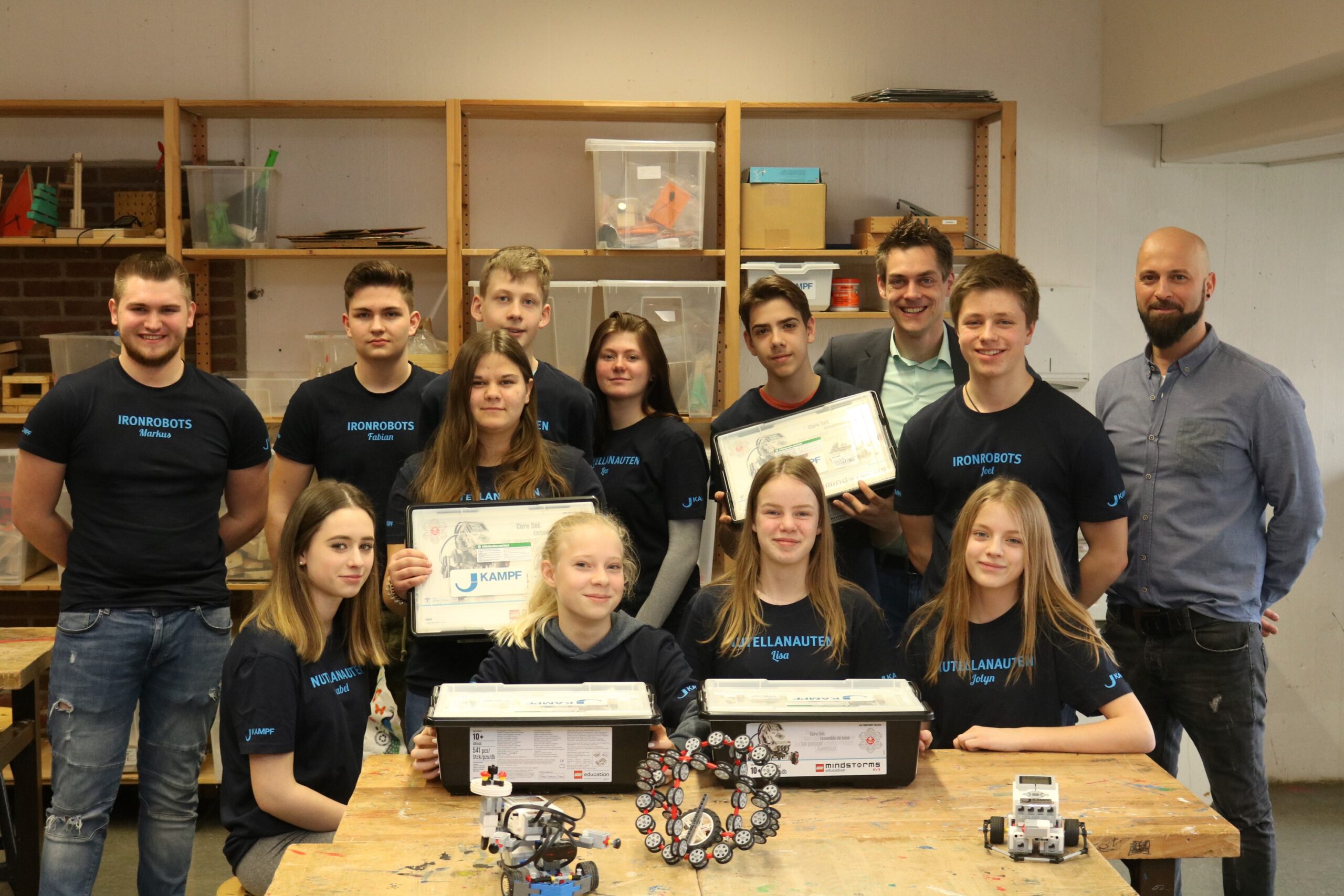 Gruppe von Schülern mit Urkunden und Robotern in einer Werkstatt.