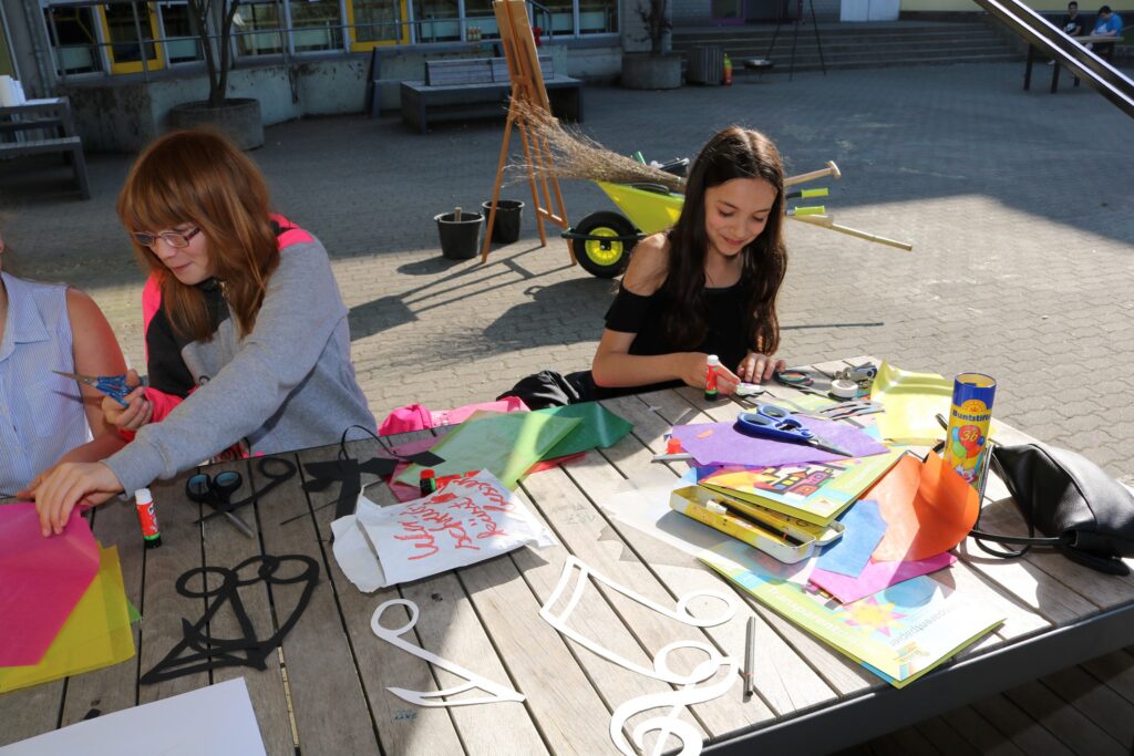 Schülerinnen basteln mit buntem Papier und Scheren an einem Tisch im Freien.