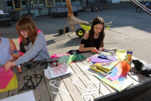 Schülerinnen basteln mit buntem Papier und Scheren an einem Tisch im Freien.