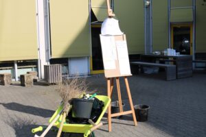 Gartengeräte und eine Staffelei stehen im Freien vor einem Gebäude.