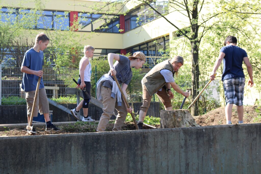 Sechs Menschen arbeiten mit Schaufeln im Freien an einem Hang. Hintergrund: Schule.