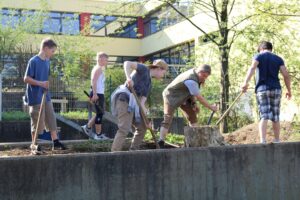 Sechs Menschen arbeiten mit Schaufeln im Freien an einem Hang. Hintergrund: Schule.