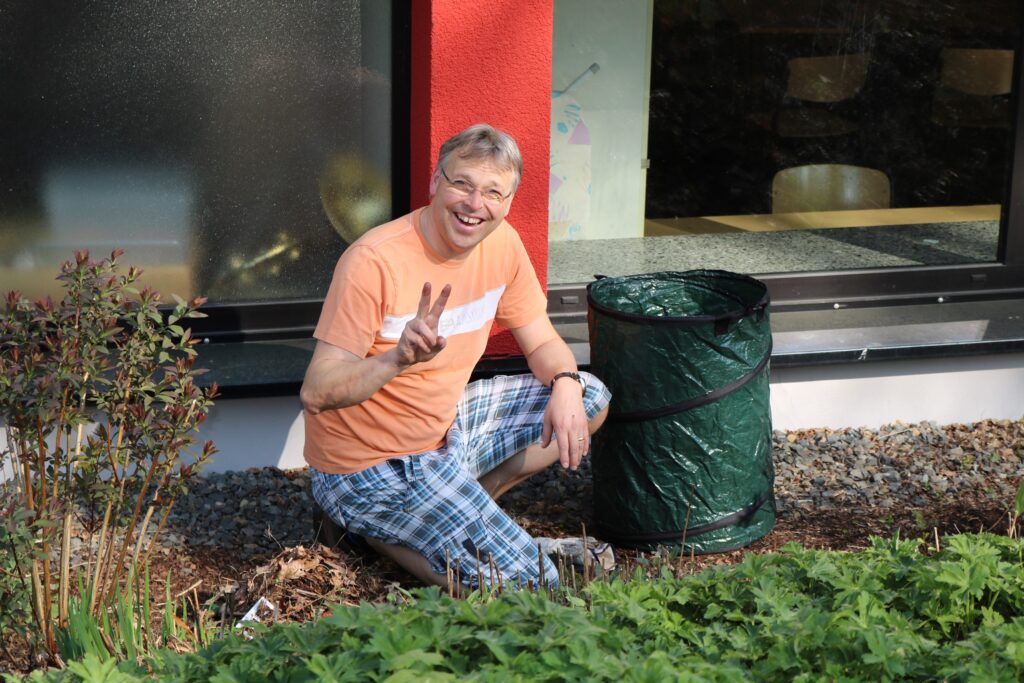 Mann kniet im Garten neben einem Abfallbehälter und zeigt das Peace-Zeichen.