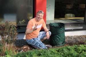 Mann kniet im Garten neben einem Abfallbehälter und zeigt das Peace-Zeichen.