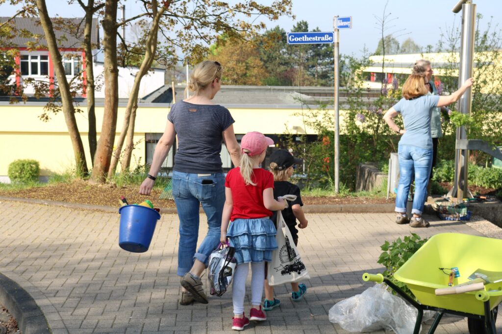 Frau mit Kind und Eimer geht auf einem Pflasterweg, im Hintergrund eine andere Person.