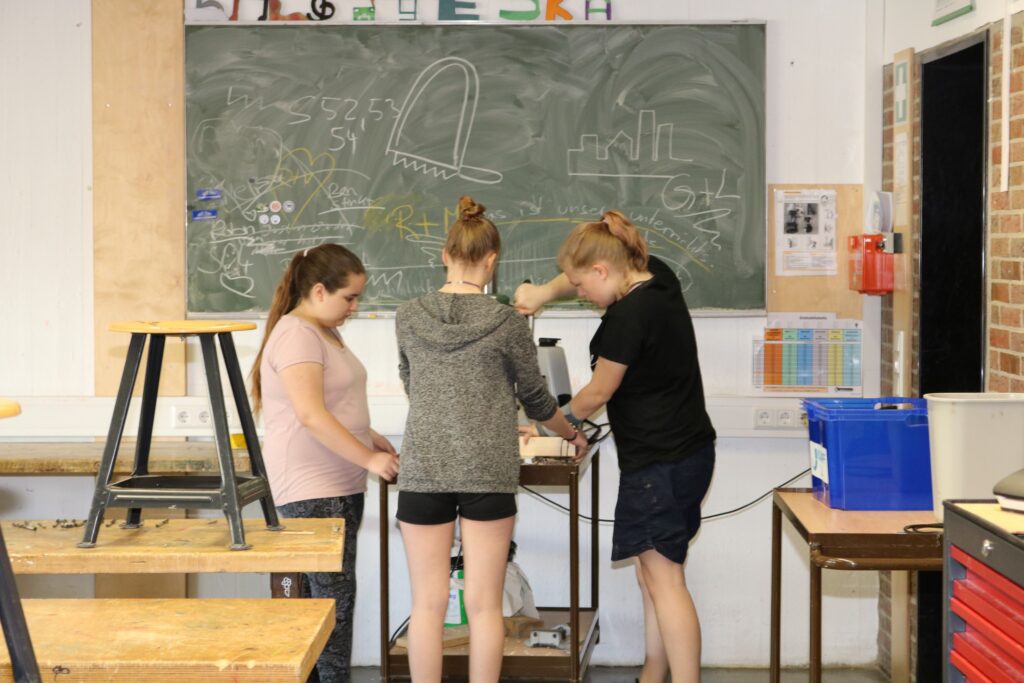 Drei Schülerinnen arbeiten gemeinsam an einem Projekt in einer Werkstatt.