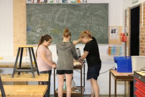 Drei Schülerinnen arbeiten gemeinsam an einem Projekt in einer Werkstatt.