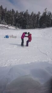 Zwei Kinder in warmer Kleidung stehen auf Schnee und helfen sich beim Skifahren.