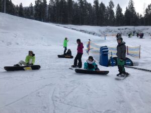 Schüler in Skikleidung sitzen auf Snowboards im Schnee. Wälder im Hintergrund.