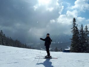 Person auf Skiern steht im verschneiten Gebirge. Wolken und Bäume im Hintergrund.
